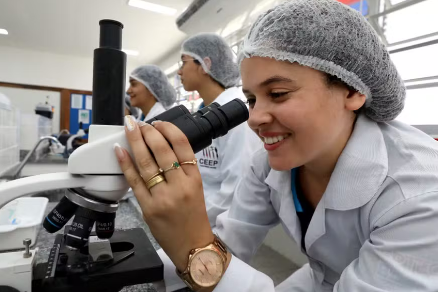 A imagem mostra uma mulher de pele branca usando touca e jaleco em um laboratório. Ela esta olhando uma lâmina através do microscópio