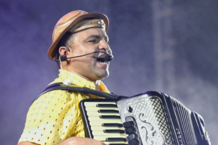O cantor chambinho do acordeom esta cantando enquanto toca uma sanfona. ele usa chapéu de cangaceiro